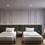 A bright, luxurious twin stateroom on the Hansa Spirit Nile Cruise featuring two single beds with plush green velvet upholstered headboards and elegant white bedding with light green accents. The wall behind the beds is grey with classical wall molding. To the right, two stylish, curved salmon-colored velvet chairs sit by a window with sheer and grey blackout curtains. The floor is light wood, and abstract artwork is on the left wall.