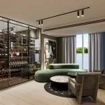 A luxurious, modern lounge space featuring a glass-enclosed, floor-to-ceiling wine cellar, a curved green velvet sofa, a round marble coffee table on a patterned rug, and two upholstered armchairs. Large windows with grey curtains offer a view of an outdoor waterway or lake. The room has light wood floors and black track lighting.