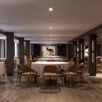 A spacious and elegant main dining room on the Hansa Spirit Nile cruise ship. The room features multiple round tables covered with light tablecloths and surrounded by warm, brown leather chairs. Dark wood columns frame the space, and the floor is polished wood. Abstract art hangs on a back wall, and soft lighting illuminates the room.