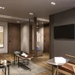 A modern, luxurious sitting area or lounge within a stateroom on the Hansa Spirit Nile Cruise. The space features light wood floors, an armchair upholstered in a reddish-brown velvet, a white accent chair, and a two-tiered oval coffee table on a grey rug. The wall is dark grey with a mounted TV, and the room has several doors with dark wood finishes and a partial view into a dressing area.