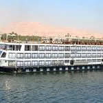 A large, white and black multi-deck Nile cruise ship named "Hansa Egypt" on a blue river under a bright, clear sky. The ship has rows of windows across its entire side and a shaded sundeck on the top level. The background features lush green vegetation, buildings along the riverbank, and arid, tawny-colored hills under a hazy sky.