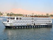 A large, white and black multi-deck Nile cruise ship named "Hansa Egypt" on a blue river under a bright, clear sky. The ship has rows of windows across its entire side and a shaded sundeck on the top level. The background features lush green vegetation, buildings along the riverbank, and arid, tawny-colored hills under a hazy sky.