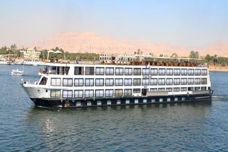 A Large, White And Black Multi-Deck Nile Cruise Ship Named &Amp;Quot;Hansa Egypt&Amp;Quot; On A Blue River Under A Bright, Clear Sky. The Ship Has Rows Of Windows Across Its Entire Side And A Shaded Sundeck On The Top Level. The Background Features Lush Green Vegetation, Buildings Along The Riverbank, And Arid, Tawny-Colored Hills Under A Hazy Sky.