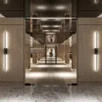 A modern, luxurious Stateroom on the Hansa Spirit Nile Cruise featuring a large bed with a deep green velvet upholstered headboard and crisp white bedding. A partial dark wood and black frame partition separates the sleeping area from a living/dressing area, incorporating a wall-mounted TV and a floating vanity desk with two round, green velvet stools. The decor includes abstract, mid-century modern style artwork.