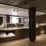 A modern, upscale buffet area on the Hansa Spirit Nile cruise ship. The central counter has a curved design with vertical wood slat paneling, marble countertops, and stacked plates visible on a shelf below. Several covered chafing dishes and serving bowls are set out. The room features dark wood columns, matching cabinetry, and large windows with natural light in the background.