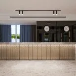 A sleek, modern reception desk with a light wood paneled front and a grey marble countertop. Behind the desk, a dark, reflective panel features three clocks displaying different time zones. To the left, large windows with blue-grey curtains offer a bright view of a waterfront or outdoor landscape. The floor is light grey marble, and the ceiling has black track lighting.