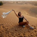 Happy-smiling-blonde-woman-sit-in-desert-sand-with-fluttering-on-wind-white-shirt-enjoy-safari-tour-in-Dubai