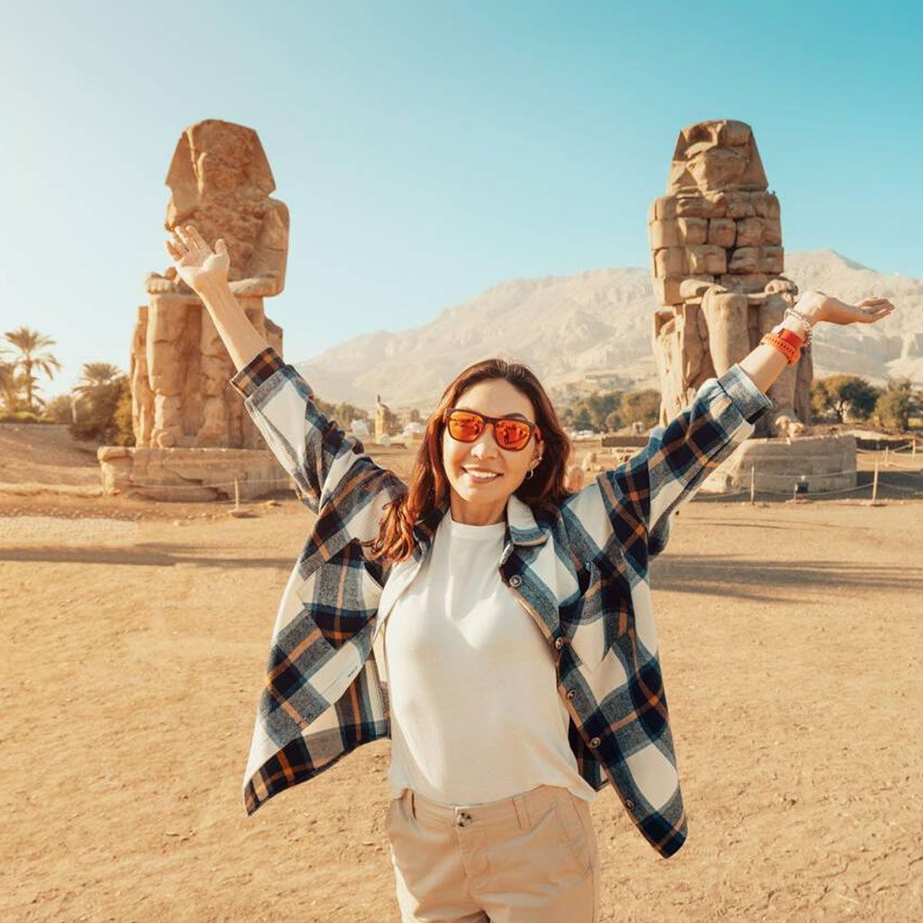 A happy tourist girl taking a photo with the Colossi of Memnon during her 8 day Egypt tour package, arms raised in excitement before the ancient statues.