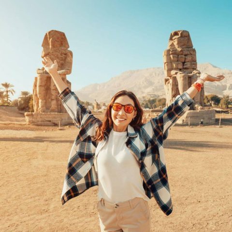 A happy tourist girl taking a photo with the Colossi of Memnon during her 8 day Egypt tour package, arms raised in excitement before the ancient statues.