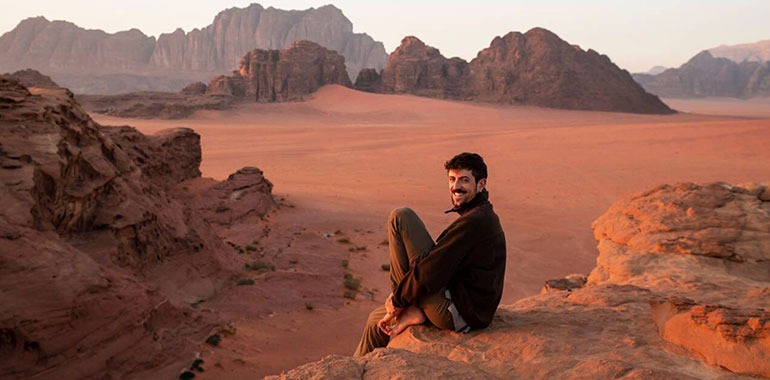 Happy Young Man Sitting On Rock In The Desert At Sunset