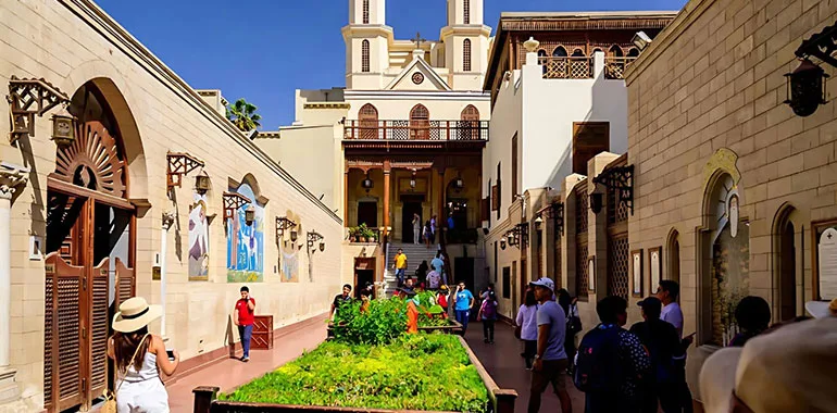 A Lively, Sunlit View Of The Courtyard And Entrance Of The Hanging Church In Coptic Cairo, With Tourists And Visitors Walking Toward The Ancient Building With Its Wooden Balcony And Bell Towers.