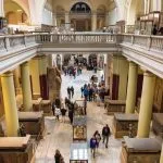 Tourists explore the vast central hall and upper balconies of the Inside the Egyptian museum of Cairo, with many large ancient Egyptian sarcophagi and statues on display.