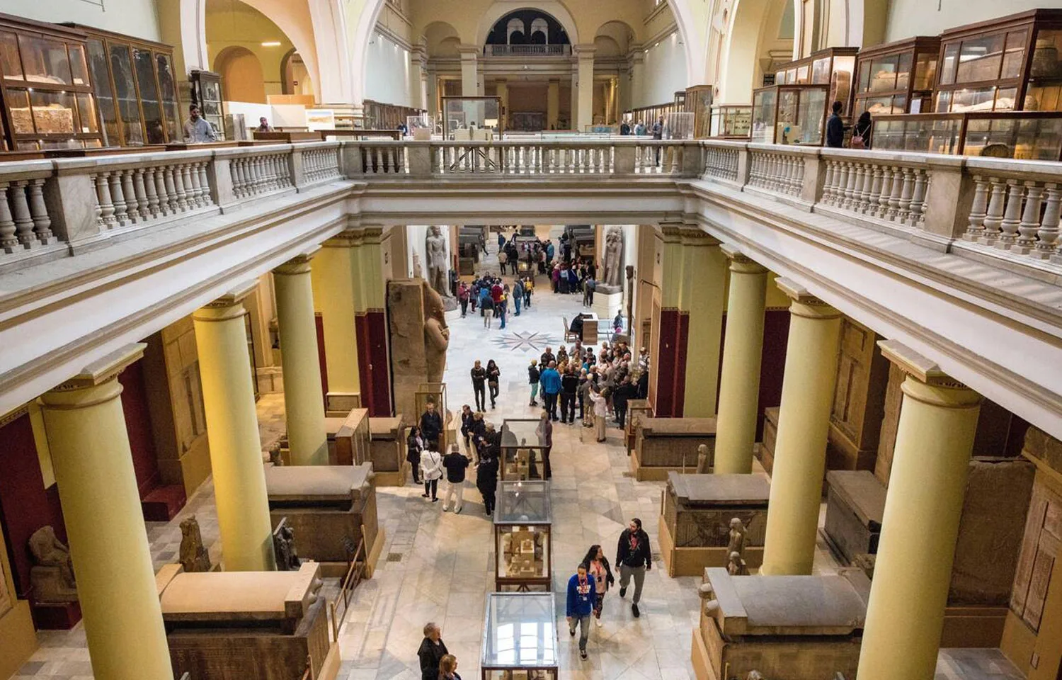 Tourists explore the vast central hall and upper balconies of the Inside the Egyptian museum of Cairo, with many large ancient Egyptian sarcophagi and statues on display.