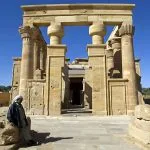 The large, well-preserved sandstone entrance gateway and columns of the Kharga Oasis Temple Of Hibis Egypt, framed by a clear blue sky and a bedouin figure resting nearby.