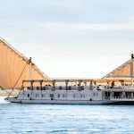 A traditional two-masted wooden sailing boat, known as a Dahabiya, glides along the wide Nile River under a bright sky with lush green banks visible in the distance.
