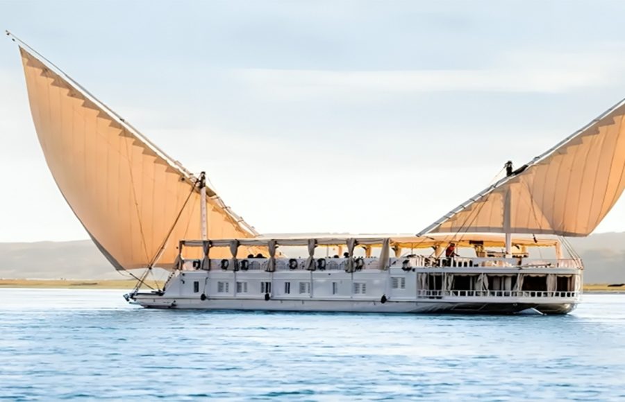 A traditional two-masted wooden sailing boat, known as a Dahabiya, glides along the wide Nile River under a bright sky with lush green banks visible in the distance.