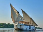A traditional two-masted wooden sailing boat, known as a Dahabiya, glides along the wide Nile River under a bright sky with lush green banks visible in the distance.