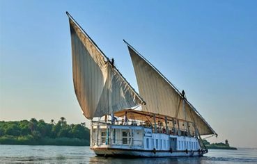 A traditional two-masted wooden sailing boat, known as a Dahabiya, glides along the wide Nile River under a bright sky with lush green banks visible in the distance.