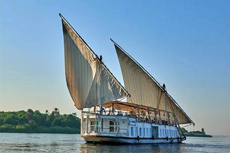 A traditional two-masted wooden sailing boat, known as a Dahabiya, glides along the wide Nile River under a bright sky with lush green banks visible in the distance.