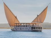 A traditional Egyptian two-masted wooden sailing boat (dahabiya) named Lazuli Albatros cruising on the Nile river with lush green banks in the background.