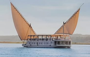 A traditional Egyptian two-masted wooden sailing boat (dahabiya) named Lazuli Albatros cruising on the Nile river with lush green banks in the background.