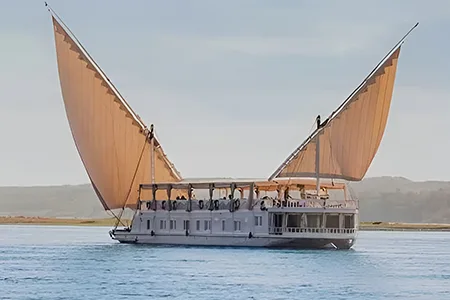 A traditional Egyptian two-masted wooden sailing boat (dahabiya) named Lazuli Albatros cruising on the Nile river with lush green banks in the background.