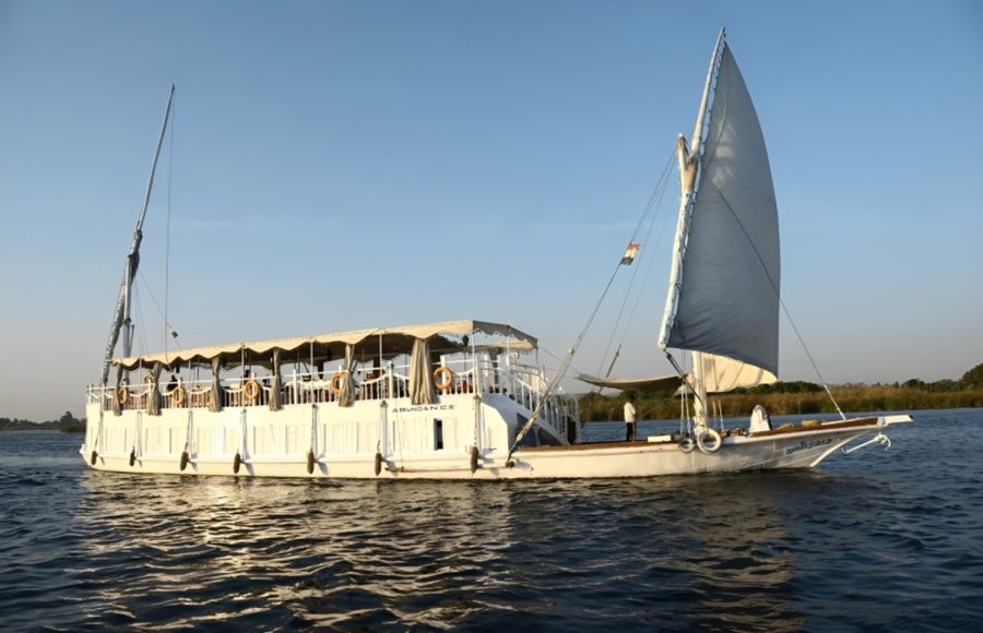 A traditional, two-masted wooden sailing boat, known as a Dahabiya, cruising on the Nile River with white sails full of wind.