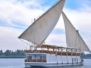 A traditional, two-masted wooden sailing boat, known as a Dahabiya, cruising on the Nile River with white sails full of wind.