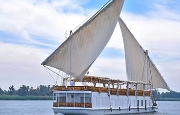 A traditional, two-masted wooden sailing boat, known as a Dahabiya, cruising on the Nile River with white sails full of wind.