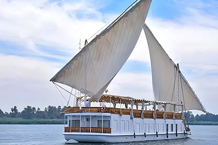 A traditional, two-masted wooden sailing boat, known as a Dahabiya, cruising on the Nile River with white sails full of wind.