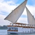 A traditional, two-masted wooden sailing boat, known as a Dahabiya, cruising on the Nile River with white sails full of wind.