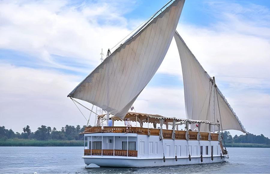 A traditional, two-masted wooden sailing boat, known as a Dahabiya, cruising on the Nile River with white sails full of wind.