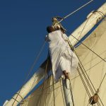 A crew member in a white galabeya climbing the wooden mast of the ship.