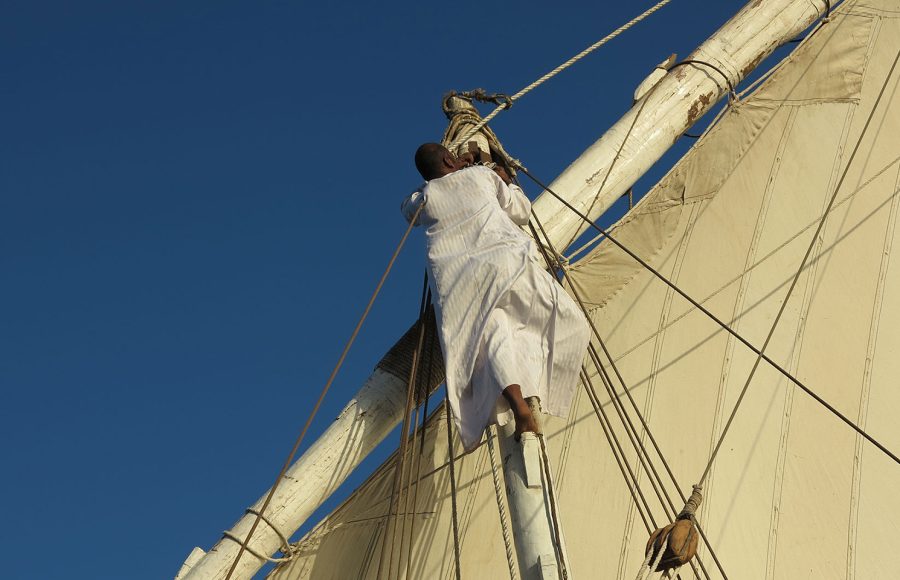 A crew member in a white galabeya climbing the wooden mast of the ship.