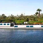 Full length of the Merit Star Dahabiya boat on the water with palm trees and green fields in the background.