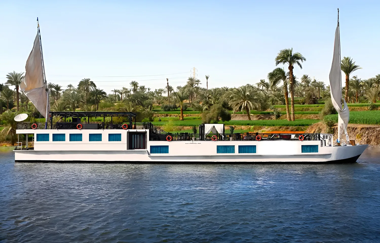 Full length of the Merit Star Dahabiya boat on the water with palm trees and green fields in the background.