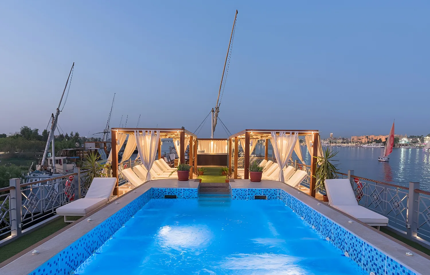 Nighttime view of a lit swimming pool on the deck of a Dahabiya, flanked by white cabanas and glowing lights.