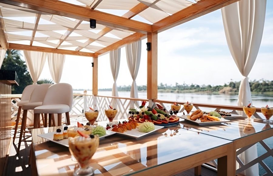 A spread of fresh fruit and desserts on a glass table under a white canopy on a boat deck.