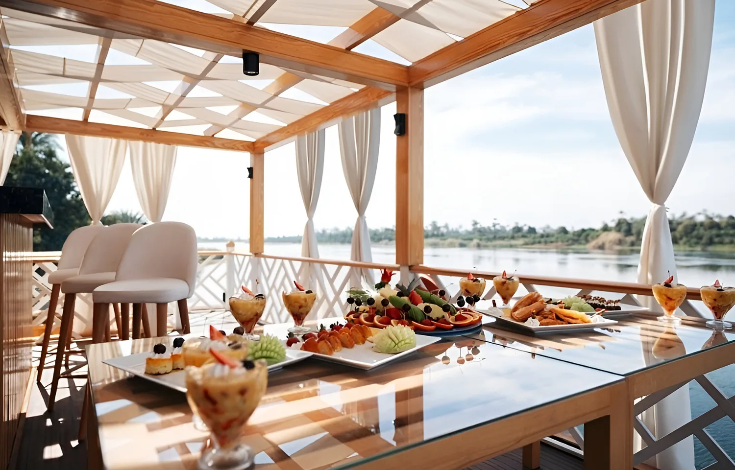 A spread of fresh fruit and desserts on a glass table under a white canopy on a boat deck.