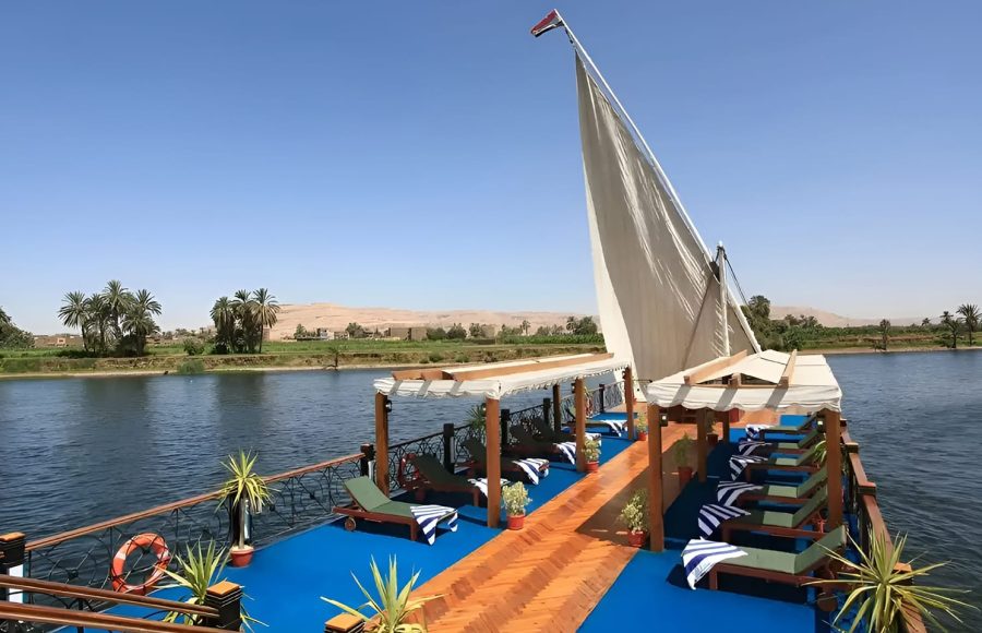 Wide view of the blue-carpeted sundeck of the Merit Star Dahabiya with a large white sail against a desert landscape.