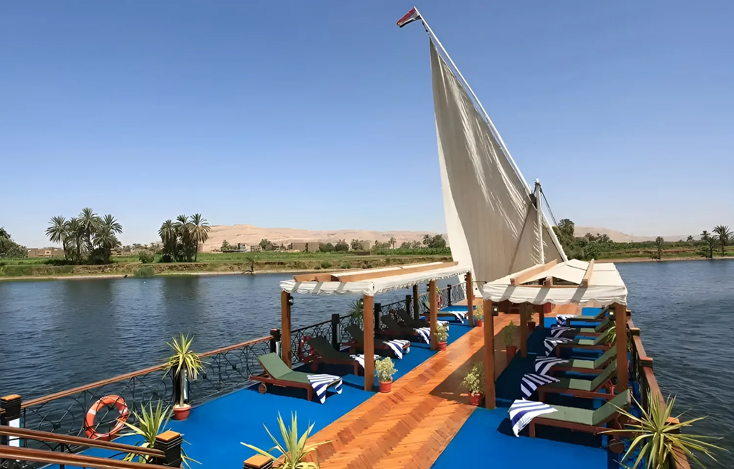 Wide view of the blue-carpeted sundeck of the Merit Star Dahabiya with a large white sail against a desert landscape.