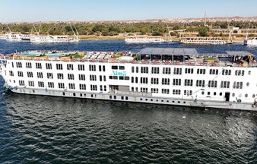 The white deluxe MS Alma Nile Cruise sailing along the Nile River, featuring an expansive sundeck with a swimming pool and panoramic views of the verdant Egyptian riverbank.