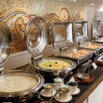 A delicious spread at the open buffet aboard the MS Alma Nile Cruise, featuring shiny chafing dishes filled with warm meals and a stunning mosaic wall backdrop.