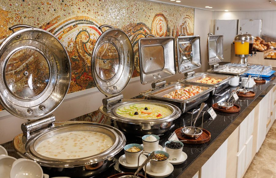 A delicious spread at the open buffet aboard the MS Alma Nile Cruise, featuring shiny chafing dishes filled with warm meals and a stunning mosaic wall backdrop.