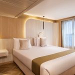 A modern and stylish Junior Suite aboard the MS Alma Nile Cruise, featuring a comfortable double bed, warm wood paneling, and a large window with sheers overlooking the river.
