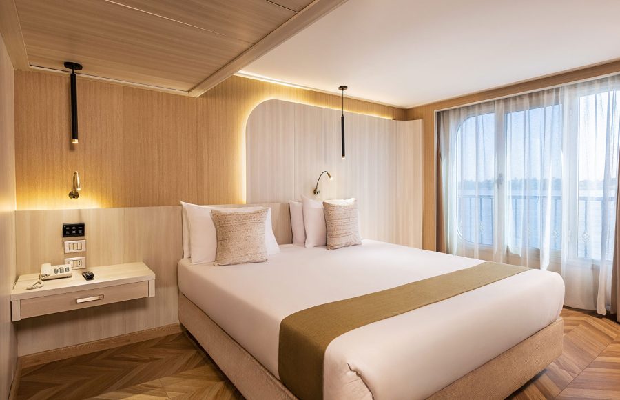 A modern and stylish Junior Suite aboard the MS Alma Nile Cruise, featuring a comfortable double bed, warm wood paneling, and a large window with sheers overlooking the river.