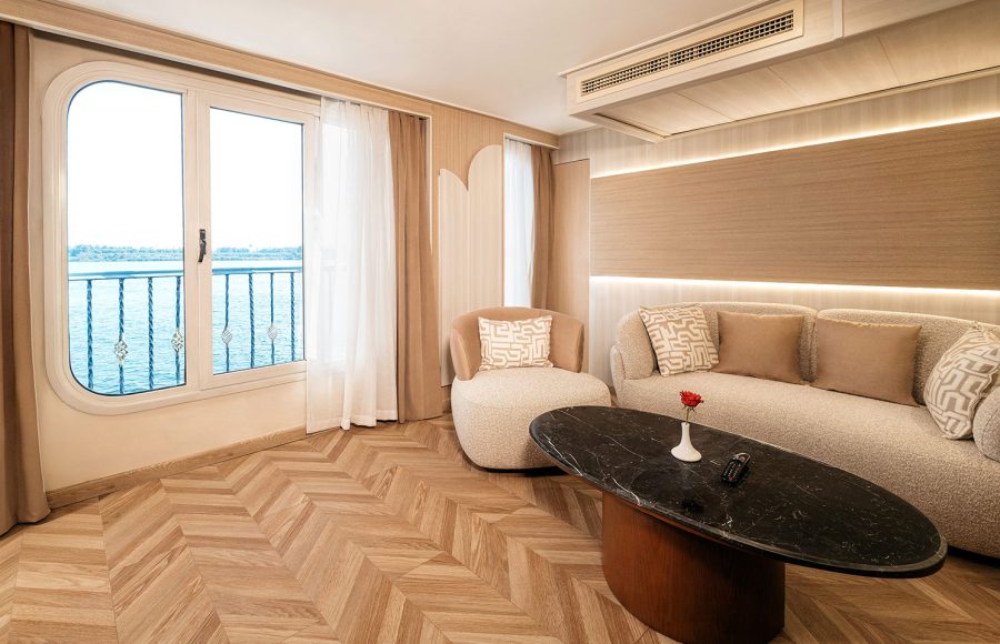 The elegant living area of the presidential suite on the MS Alma Nile Cruise, featuring a modern sofa, a stylish coffee table, and a large window with panoramic views of the river.