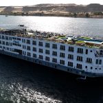 The MS Alma Nile Cruise vessel sailing smoothly along the waters of the Nile River, with desert banks and palm trees visible in the background.