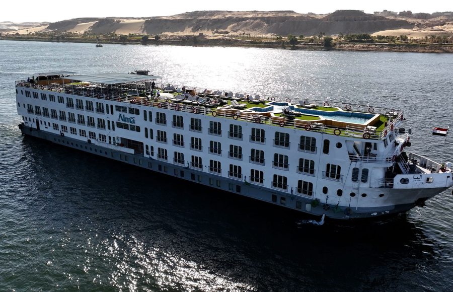 The MS Alma Nile Cruise vessel sailing smoothly along the waters of the Nile River, with desert banks and palm trees visible in the background.