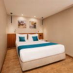 A cozy, modern Standard Cabin aboard the MS Alma Nile Cruise, featuring a comfortable double bed dressed in white and teal linens, warm wood flooring, and elegant bedside lighting.